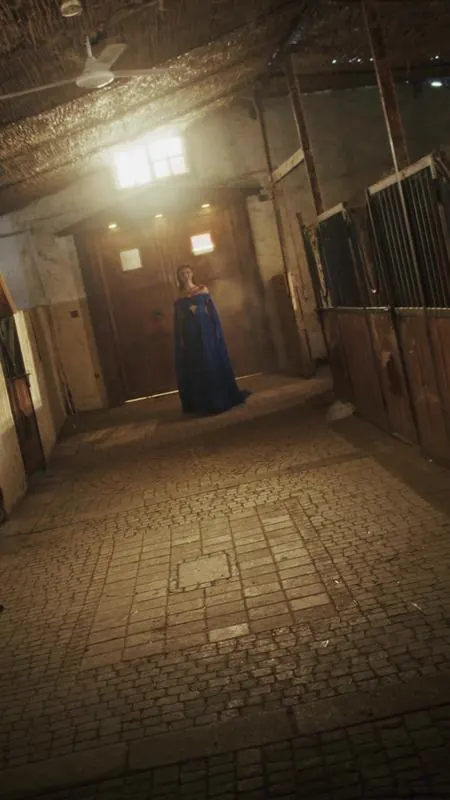 Lead dancer in a blue gown at the end of a lit stable corridor